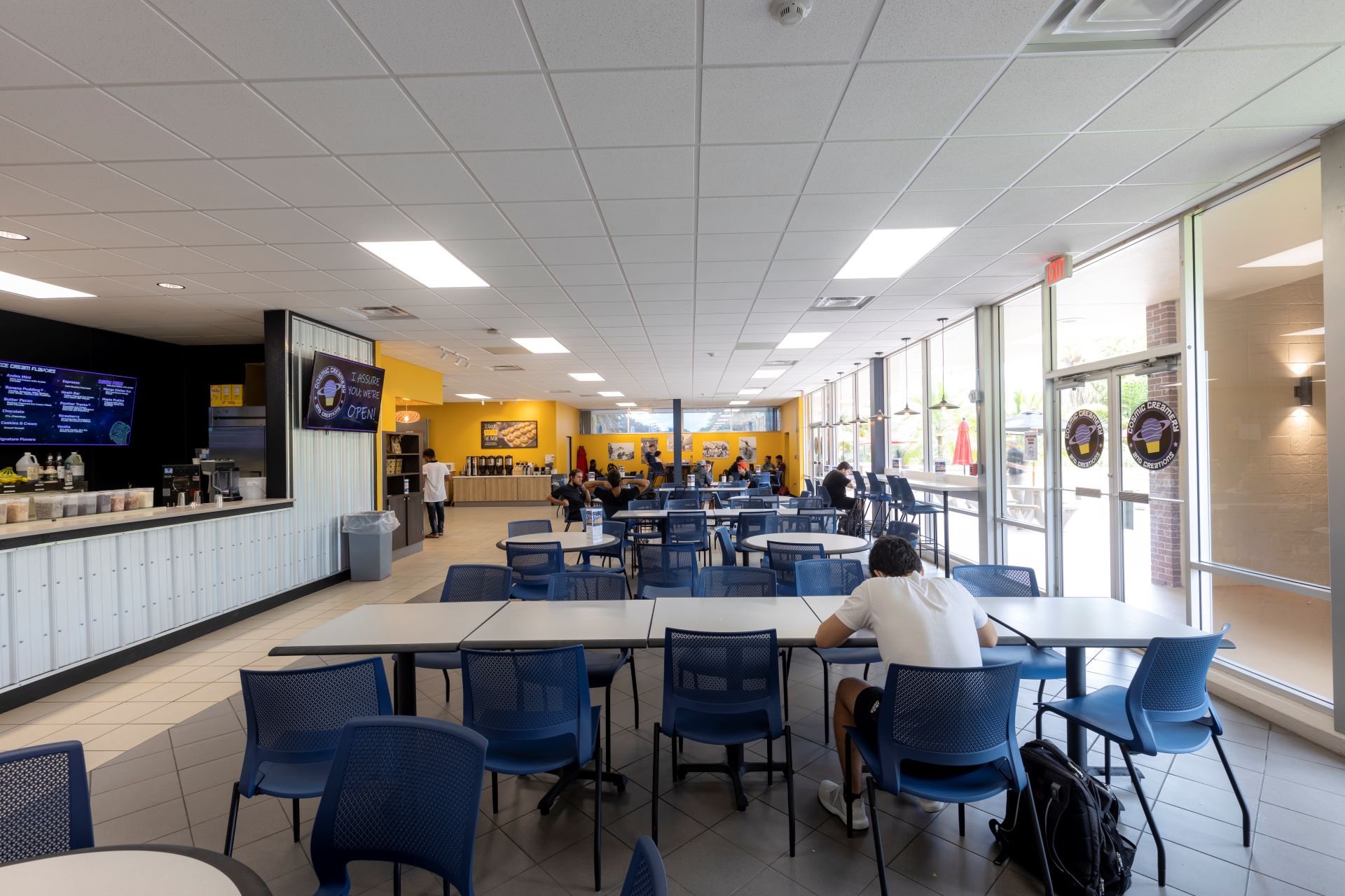 A student cafeteria with blue chairs and wooden tables, occasionally occupied by students